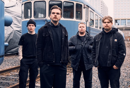 Neljä muusikkoa seisoo junavarikkoalueella vanhan junavaunun edessä.